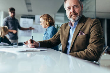 Mature businessman sitting in conference room with colleagues in background. Senior man sitting in board room during presentation. - JLPSF02555