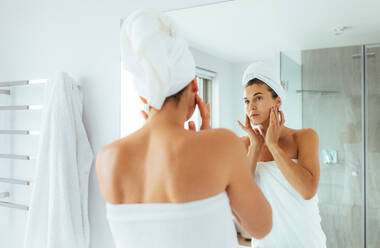 Woman observing her face in mirror in bathroom. Woman in towels wrapped around head and body after bath. - JLPSF02559