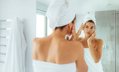 Woman massaging her face looking at a mirror in bathroom. Woman in towels wrapped around head and body after bath. - JLPSF02560