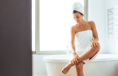 Frau trägt nach dem Bad Feuchtigkeitscreme auf ihre Beine auf. Frau mit Handtüchern um den Kopf gewickelt sitzt auf der Badewanne im Badezimmer. - JLPSF02562