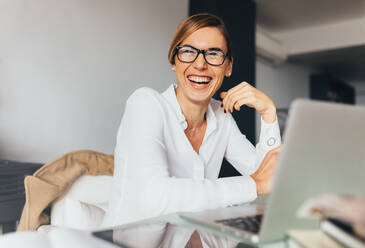 Geschäftsfrau mit Brille sitzt an ihrem Schreibtisch im Büro und lacht. Frau sitzt im Büro mit Laptop-Computer auf ihrem Schreibtisch. - JLPSF02578