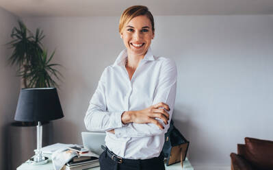 Smiling business woman standing with folded arms in office. Woman in formal business attire standing in front of her desk in office. - JLPSF02582
