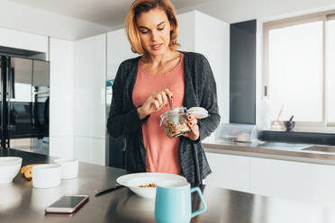 Frau nimmt Haferflocken aus einem Glasgefäß auf einen Teller. Frau bereitet ein gesundes Frühstück in ihrer Küche vor. - JLPSF02583