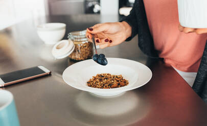 Woman preparing a healthy breakfast in her kitchen. Woman adding blueberry toppings to her oatmeal. - JLPSF02586