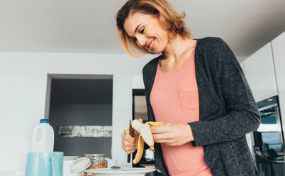 Frau, die in ihrer Küche ein gesundes Frühstück zubereitet. Lächelnde Frau, die Bananenscheiben auf ihren Teller mit Haferflocken legt. - JLPSF02587