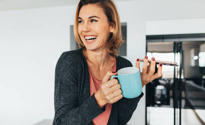 Woman talking over mobile phone on loudspeaker holding a coffee mug. Smiling woman enjoying coffee while talking over phone at home. - JLPSF02589