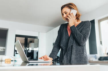 Frau, die zu Hause über ihr Mobiltelefon spricht und ihren Laptop bedient. Geschäftsfrau, die sich am Frühstückstisch mit Büroarbeit beschäftigt. - JLPSF02590