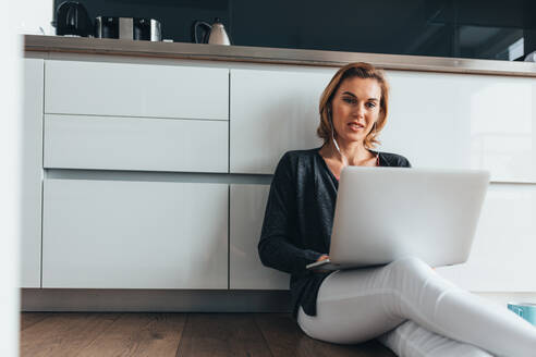 Frau mit Laptop-Computer auf dem Küchenboden sitzend und mit Kopfhörern. - JLPSF02595