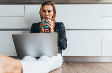 Woman sitting on floor holding a coffee mug working on laptop computer. Woman using laptop computer wearing earphones. - JLPSF02598