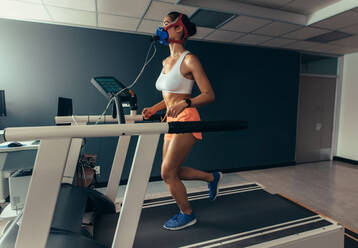 Female runner with mask running on treadmill machine testing her performance. Woman athlete examining her fitness in biomechanics lab. - JLPSF02625