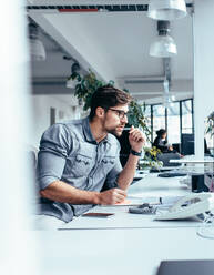 Vertikale Aufnahme eines nachdenklichen Geschäftsmannes, der auf einen Monitor schaut. Ein junger Mann sitzt im Büro und denkt über seine Arbeit nach. - JLPSF02643