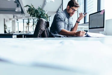 Geschäftsmann, der mit seinem Handy telefoniert und sich Dokumente ansieht. Junge männliche Führungskraft bei der Arbeit im Büro. - JLPSF02646