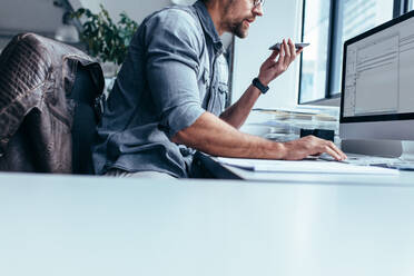 Businessman with mobile phone sitting in office and working on computer. Young businessman talking on mobile phone and using computer in office. - JLPSF02647