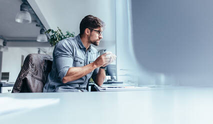 Businessman sitting in office with cup of coffee. Young man holding mug and looking away. - JLPSF02649