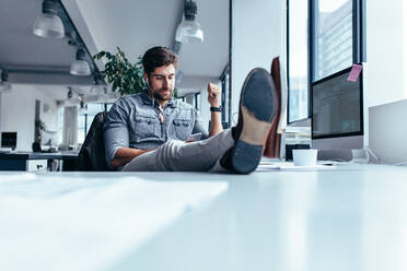 Junger Mann hört Musik am Telefon am Arbeitsplatz mit den Füßen auf dem Tisch. Geschäftsmann entspannt im Büro während der Pause. - JLPSF02652
