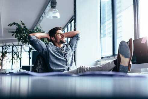 Junger Mann, der sich ausruht und Musik hört, während er im Büro sitzt. Männlicher Designer sitzt entspannt an seinem Arbeitsplatz. - JLPSF02656