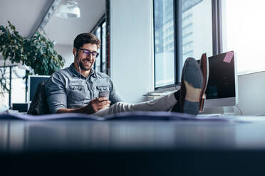 Geschäftsmann mit Handy im Büro mit den Füßen auf dem Tisch. Männlicher Designer hört Musik in der Pause. - JLPSF02657