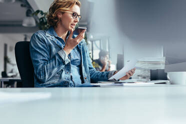 Junge Geschäftsfrau im Gespräch über Lautsprecher mit Dokument. Junge Frau sitzt im Büro mit Smartphone und schaut auf ein Papier. - JLPSF02662