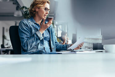 Young businesswoman talking on speaker phone with document. Young woman sitting in office with smart phone and looking at a paper. - JLPSF02662