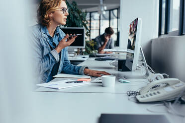 Junge Geschäftsfrau, die über Lautsprecher telefoniert und am Computer arbeitet. Junge Frau sitzt vor einem Monitor mit Smartphone. - JLPSF02665