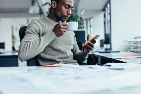 Businessman sitting at office desk having a coffee. Young african man with cup of coffee and smart phone. - JLPSF02676