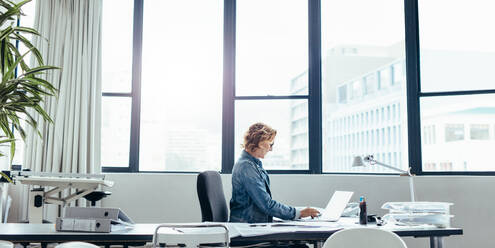 Side view of female executive sitting in her office using laptop. Young caucasian woman looking busy working on laptop. - JLPSF02679