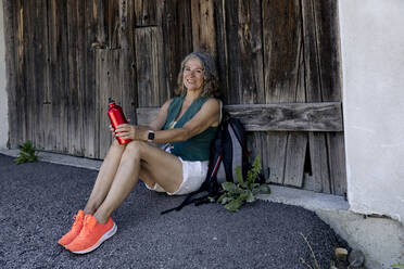 Lächelnde ältere Frau, die eine Wasserflasche hält und sich an eine Holztür lehnt - FLLF00752