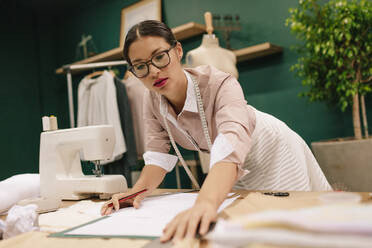 Asiatische Modedesignerin bei der Anfertigung einer Design-Skizze auf Papier. Schneiderin bei der Arbeit an neuen Kleidungsentwürfen in ihrer Werkstatt. - JLPSF02697