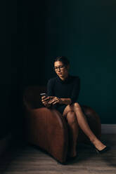 Sophisticated woman using a mobile phone while sitting on an armchair in her office. Modern woman reading a text message on her smartphone. - JLPSF02698