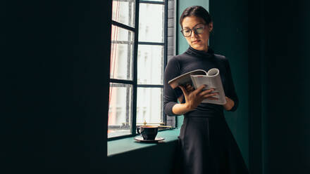 Asiatische Frau steht am Fenster und liest ein Buch. Schöne Frau liest eine Modezeitschrift. - JLPSF02723