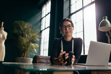 Asiatische Frau hält eine Tasse Kaffee in der Hand und blickt in die Kamera. Eine Designerin sitzt entspannt an ihrem Schreibtisch und trinkt Kaffee. - JLPSF02728