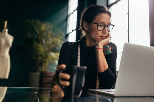Junge Modedesignerin sitzt vor einem Laptop an ihrem Arbeitstisch und recherchiert im Internet nach einem neuen Kleidungsstil. - JLPSF02730
