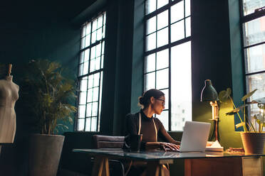 Designerin bei der Arbeit in ihrem Büro. Frau sitzt vor einem Laptop und schaut in ein Buch. - JLPSF02732