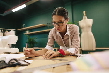 Junge asiatische Frau, die in ihrer Werkstatt arbeitet und einen Entwurf auf Papier anfertigt. Eine Modedesignerin entwirft ein neues Kleidermuster auf Papier. - JLPSF02736