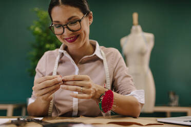 Junge asiatische Designerin beim Einfädeln der Nadel. Modedesignerin bei der Arbeit in ihrem Atelier. - JLPSF02749