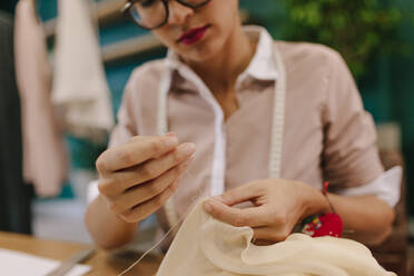 Nahaufnahme der Hände einer Frau, die an ihrem Arbeitsplatz ein Designerkleid näht. Eine Modedesignerin arbeitet in ihrem Atelier. Die Hände einer Schneiderin machen Handarbeit am Kleid. - JLPSF02752