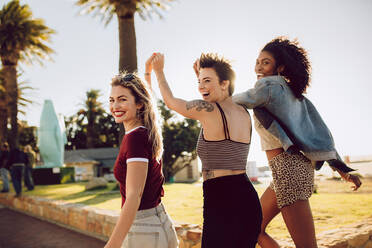 Gruppe schöner junger Frauen, die zusammen in einem Park spazieren gehen, sich an den Händen halten und lächeln. Verschiedene Freundinnen, die zusammen in einem Park Spaß haben - JLPSF02762