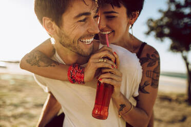 Handsome young guy piggybacking his girlfriend drinking soft drink outdoors. Happy caucasian couple enjoying themselves outdoors. - JLPSF02770