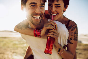 Young happy couple outdoors having fun. Man carrying his girlfriend on his back drinking soft drink from glass bottle with straw. - JLPSF02771