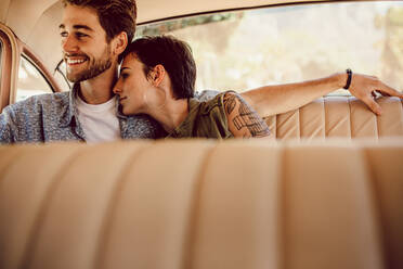Ein lächelnder junger Mann und eine lächelnde junge Frau sitzen auf dem Rücksitz eines Autos und schauen aus dem Fenster. Das Paar genießt die Reise im Auto. - JLPSF02784