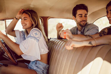 Smiling young woman driving car with her friends sitting in the backseat. Friends going on a road trip in old car. - JLPSF02788