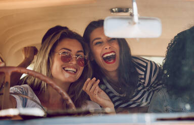 Junge Freundinnen, die in einem Auto in den Urlaub fahren. Frauen schauen in den Rückspiegel und lachen. - JLPSF02789