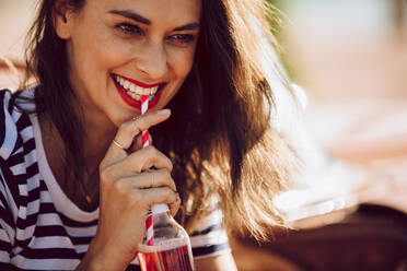Nahaufnahme einer attraktiven Frau, die ein Getränk mit Strohhalm aus einer Glasflasche trinkt. Lächelnde Frau, die an einem Sommertag ein Erfrischungsgetränk trinkt. - JLPSF02801