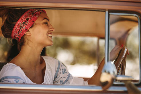 Seitenansicht einer schönen Frau mit Kopftuch, die ein Auto fährt und lächelt. Kaukasische Frau auf Road Trip mit Freunden. - JLPSF02819