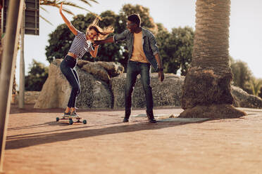 Frau lernt Longboard fahren mit Hilfe eines männlichen Freundes. Mädchen auf Skateboard mit Unterstützung eines Freundes. - JLPSF02854