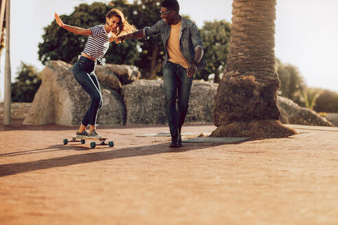 Junge Frau steht auf einem Longboard und wird von ihrem Freund unterstützt. Frau lernt im Freien Skateboard fahren. - JLPSF02855