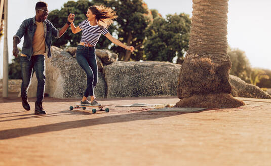 Lächelnde junge Frau auf einem Skateboard mit Unterstützung ihres Freundes. Frau beim Skaten auf dem Bürgersteig mit Freund. - JLPSF02856