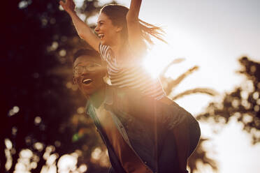 Cheerful woman with arms outstretched riding in her friends back on a sunny day. Young friends having fun outdoors. - JLPSF02859