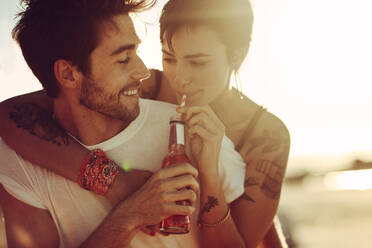 Handsome young man piggybacking his girlfriend outdoors. Woman drinking beverage while carried by her boyfriend. - JLPSF02869