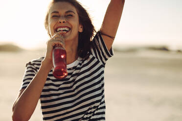 Fröhliche junge Frau steht im Freien und trinkt Cola aus einer Glasflasche. Frau genießt erfrischendes Getränk an einem heißen Sommertag. - JLPSF02870